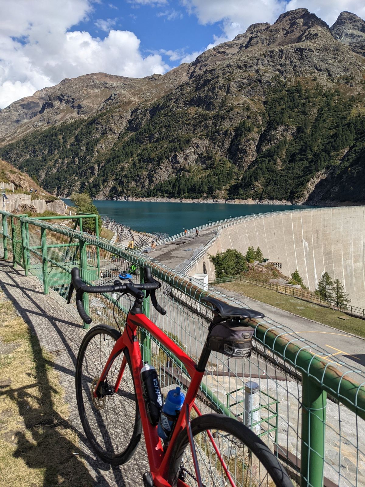 A red road bike leaning on a railing above the Place Moulin reservoir, with the dam wall and turquoise water behind
