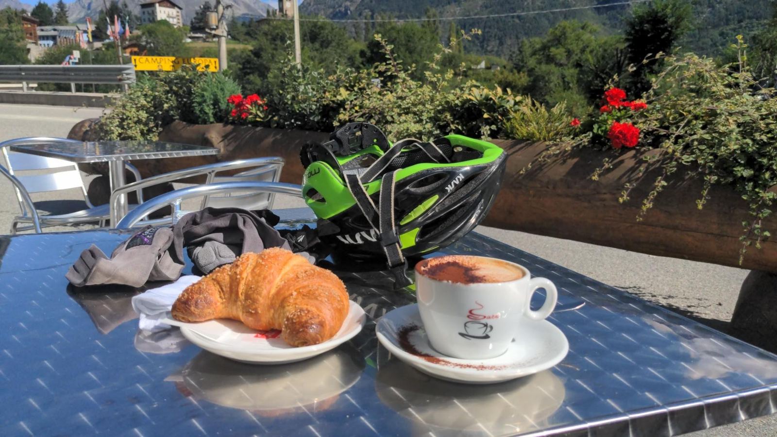 A croissant and a cup of coffee on a café table in the sun at Gignod, with a green cycling helmet and gloves