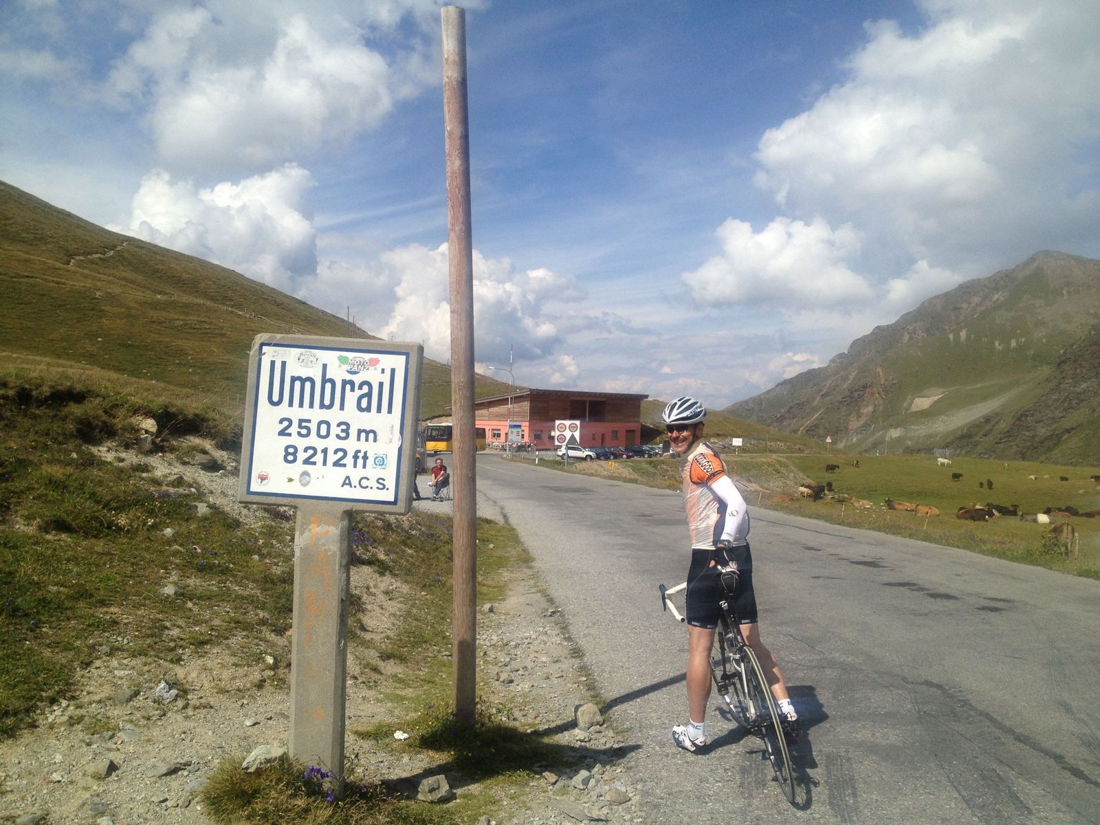 Umbrail Pass 2503m altitude sign with me and my bike, cows grazing in the background