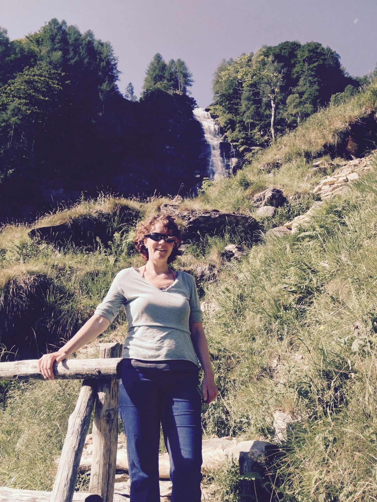 My wife standing by a wooden fence on a mountain trail near a waterfall above Sonogno